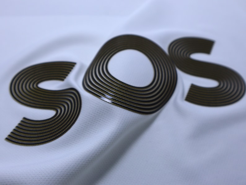 Pode incluir: Um tecido branco com a palavra "SOS" impressa em preto e dourado. As letras s&atilde;o feitas de m&uacute;ltiplos c&iacute;rculos conc&ecirc;ntricos.