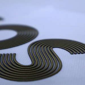 Op de afbeelding: Close-up van een witte stof met een geborduurd "S"-ontwerp in goud en zwart.