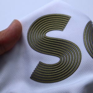 Op de afbeelding: Witte stof met een gouden, touwachtige letter "S" erop gedrukt.