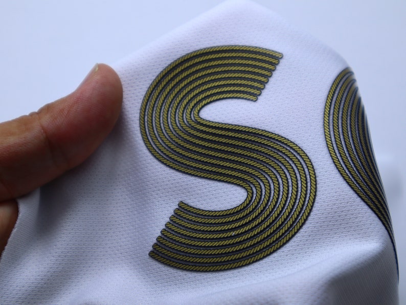 Op de afbeelding: Witte stof met een gouden, touwachtige letter "S" erop gedrukt.