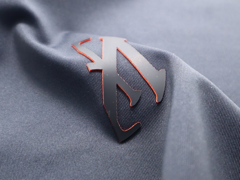 May include: A black and orange logo with a stylized letter "A" on a gray fabric background.