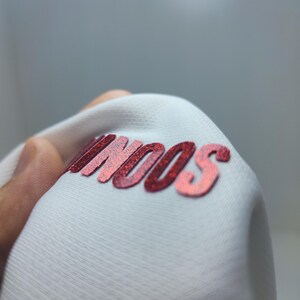 May include: Close-up of a white fabric with the word "SOON" printed in red glitter.