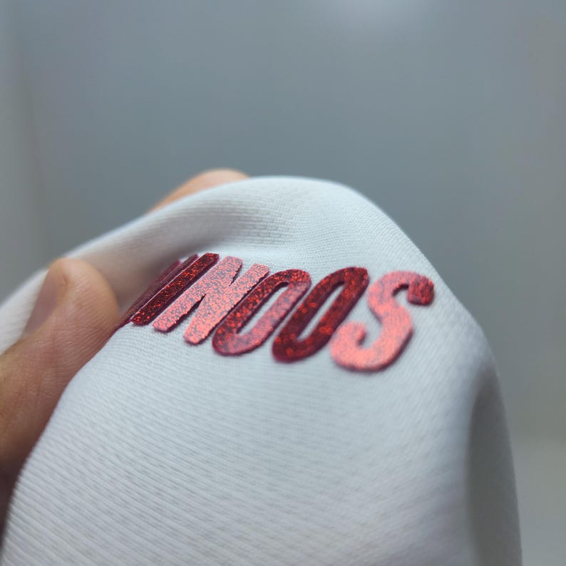 May include: Close-up of a white fabric with the word "SOON" printed in red glitter.
