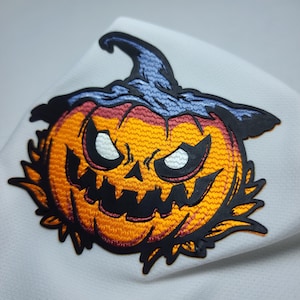 Op de afbeelding: Een zwarte en oranje geborduurde Halloween-pompoenpatch met een griezelig gezicht en een paarse hoed.