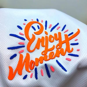 Può includere: Tessuto bianco con le parole "Enjoy Moment" in arancione, circondato da elementi decorativi blu e rosa. Il testo è in stile scritto a mano e il tessuto ha un aspetto strutturato, simile a una rete.