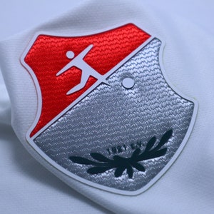 Op de afbeelding: Een witte, rode en zilveren geborduurde patch met een gestileerde figuur die een bal trapt en de tekst "1921 E.V."