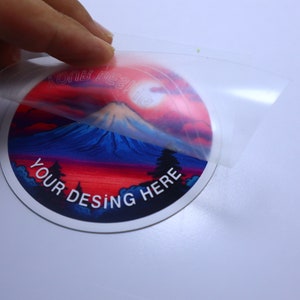K&ouml;nnte beinhalten: Ein runder Aufkleber mit einem farbenfrohen Bild einer Bergkette bei Sonnenuntergang. Der Aufkleber tr&auml;gt den Text "YOUR DESING HERE". Der Aufkleber wird von einer transparenten Schutzfolie abgezogen.