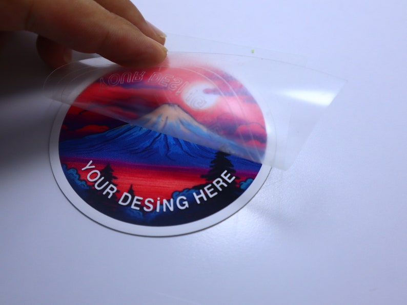 K&ouml;nnte beinhalten: Ein runder Aufkleber mit einem farbenfrohen Bild einer Bergkette bei Sonnenuntergang. Der Aufkleber tr&auml;gt den Text "YOUR DESING HERE". Der Aufkleber wird von einer transparenten Schutzfolie abgezogen.