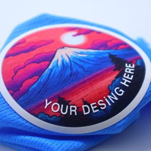 K&ouml;nnte beinhalten: Ein blauer Stoff-Patch mit wei&szlig;em Rand und einer farbenfrohen Bergszene. Der Text "YOUR DESING HERE" ist in Wei&szlig; auf dem Patch gedruckt.