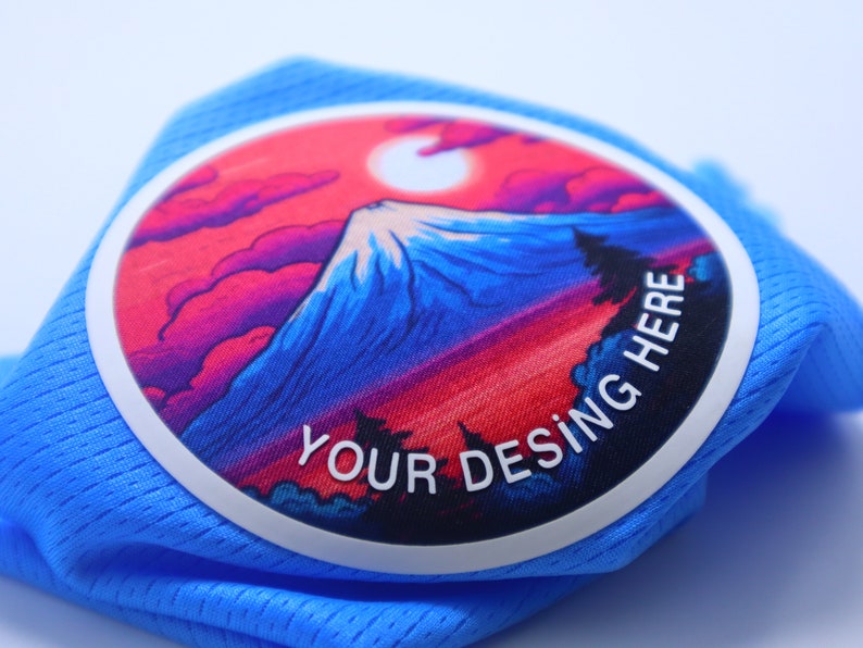 K&ouml;nnte beinhalten: Ein blauer Stoff-Patch mit wei&szlig;em Rand und einer farbenfrohen Bergszene. Der Text "YOUR DESING HERE" ist in Wei&szlig; auf dem Patch gedruckt.