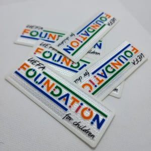 Puede incluir: Primer plano de varios parches rectangulares blancos con el texto "UEFA FOUNDATION for children" en letras azules, naranjas y verdes. Los parches tienen un fondo plateado reflectante.