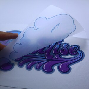 May include: A purple and blue "Paradise" design with a white backing being peeled off. The design is in a stylized font with a dripping effect.