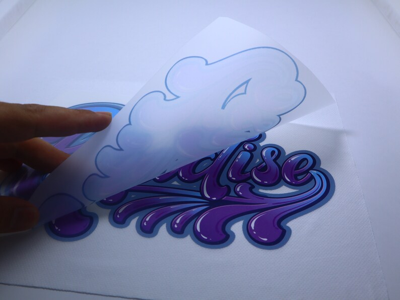 May include: A purple and blue "Paradise" design with a white backing being peeled off. The design is in a stylized font with a dripping effect.
