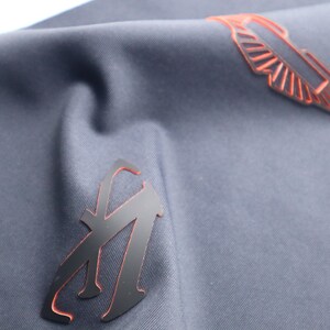 May include: A dark blue fabric with two black patches. One patch is a stylized letter "A" with an orange outline. The other patch is a stylized wing with an orange outline.