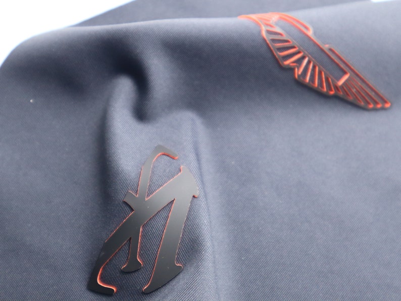 May include: A dark blue fabric with two black patches. One patch is a stylized letter "A" with an orange outline. The other patch is a stylized wing with an orange outline.