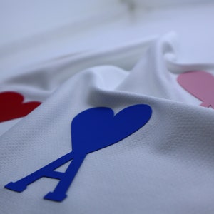 May include: White fabric with three felt hearts, one red, one pink, and one blue. The blue heart has a stylized letter A in front of it.