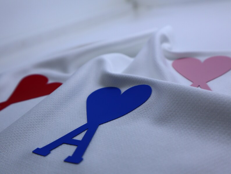 May include: White fabric with three felt hearts, one red, one pink, and one blue. The blue heart has a stylized letter A in front of it.