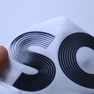 May include: Close-up of a white fabric with the letters "SO" printed in black raised lettering. The letters are made of multiple concentric circles.