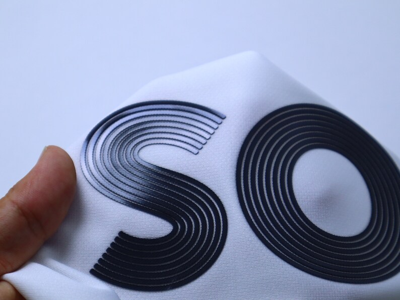May include: Close-up of a white fabric with the letters "SO" printed in black raised lettering. The letters are made of multiple concentric circles.