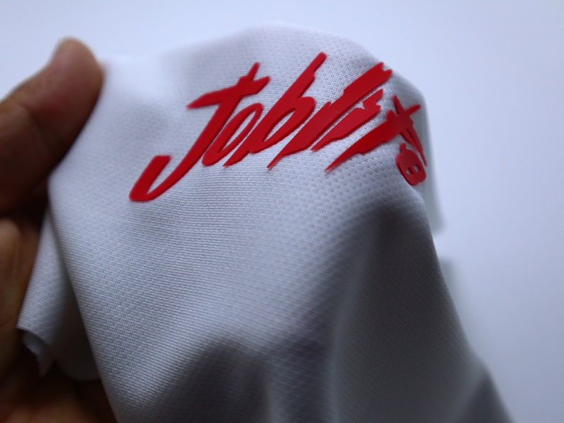 Puede incluir: Camiseta deportiva blanca con un logotipo "Joptix" rojo impreso en la parte delantera.