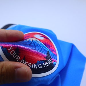 K&ouml;nnte beinhalten: Ein blauer Stoff-Patch mit einem bunten gestickten Design eines Berges und eines Sonnenuntergangs. Der Text "YOUR DESING HERE" ist in Wei&szlig; auf dem Patch gedruckt.