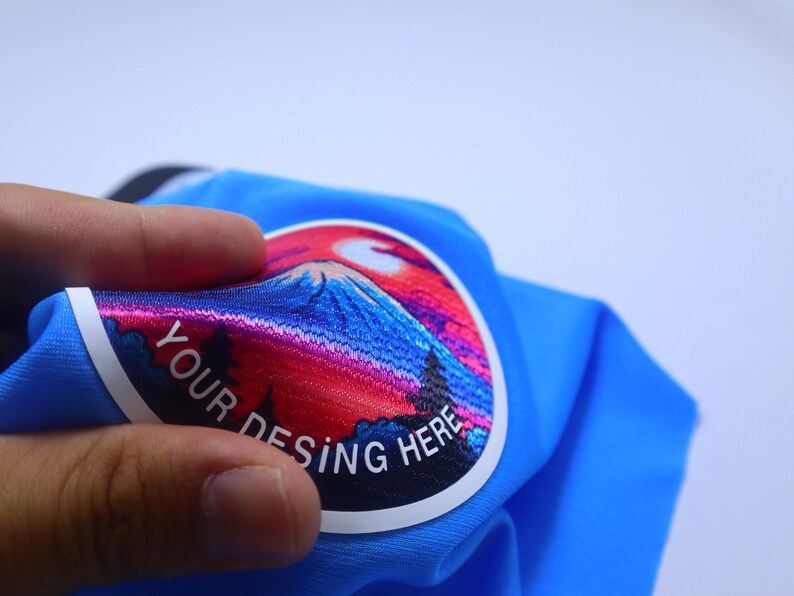 K&ouml;nnte beinhalten: Ein blauer Stoff-Patch mit einem bunten gestickten Design eines Berges und eines Sonnenuntergangs. Der Text "YOUR DESING HERE" ist in Wei&szlig; auf dem Patch gedruckt.