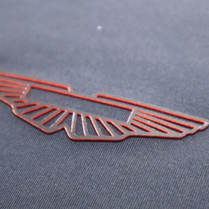 May include: A red outlined winged emblem with a stylized design. The emblem is on a dark blue fabric background.