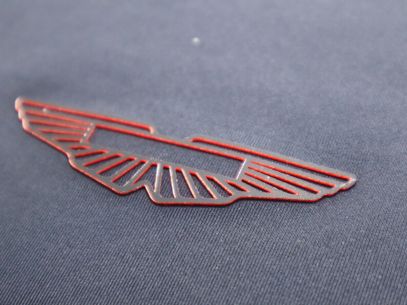 May include: A red outlined winged emblem with a stylized design. The emblem is on a dark blue fabric background.