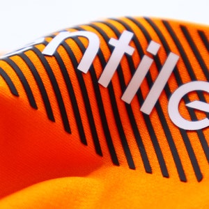 Op de afbeelding: Close-up van een feloranje stof met zwarte verticale strepen. Het woord "ntile" is in het wit gedrukt, met een verhoogd effect. De stof heeft een getextureerd oppervlak en de strepen zijn gelijkmatig verdeeld.