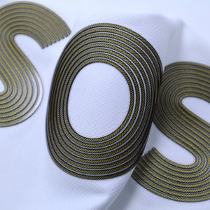 Op de afbeelding: Close-up van een witte stof met de letters "SOS" in goud en zwart. De letters zijn gemaakt van een touwachtig materiaal.