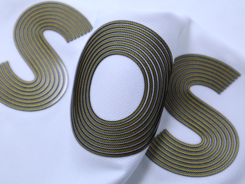 Op de afbeelding: Close-up van een witte stof met de letters "SOS" in goud en zwart. De letters zijn gemaakt van een touwachtig materiaal.