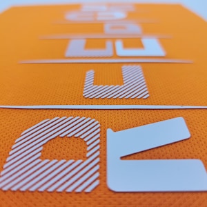 May include: Close-up of a white, raised, textured letter "U" on an orange fabric background. The letter has a striped pattern.