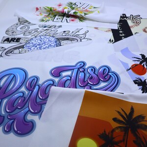 May include: Close-up of several white fabric panels with different designs printed on them. The designs include a purple and blue "Paradise" graphic, a sunset with palm trees, and a design with a planet and the words "Our Comets Are".