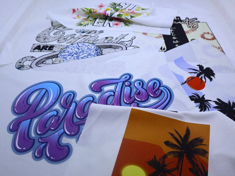 May include: Close-up of several white fabric panels with different designs printed on them. The designs include a purple and blue "Paradise" graphic, a sunset with palm trees, and a design with a planet and the words "Our Comets Are".