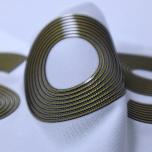 Op de afbeelding: Close-up van een witte stof met het woord "SOS" in goud gedrukt. De letters zijn gemaakt van meerdere concentrische cirkels, waardoor een textuur ontstaat.