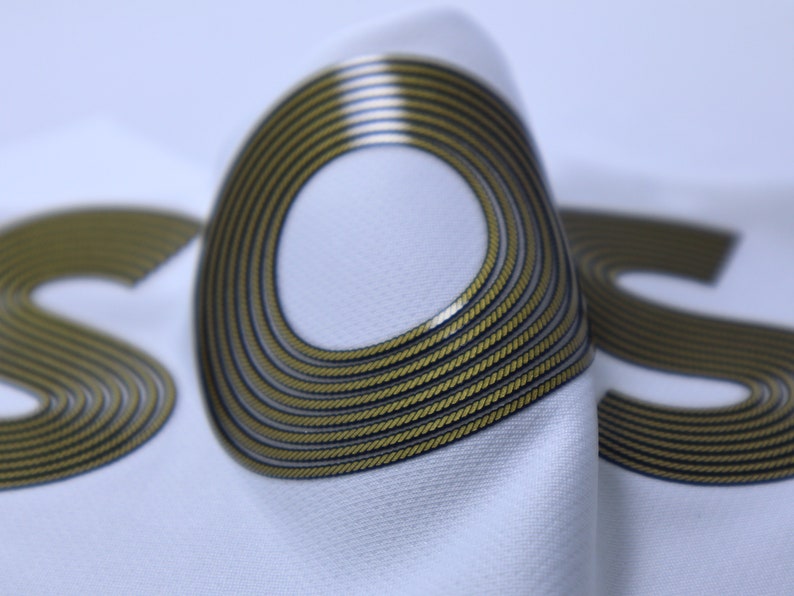 Op de afbeelding: Close-up van een witte stof met het woord "SOS" in goud gedrukt. De letters zijn gemaakt van meerdere concentrische cirkels, waardoor een textuur ontstaat.