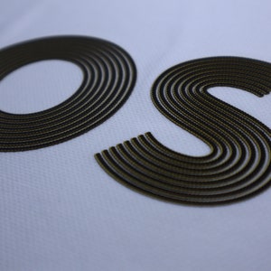 Pode incluir: Close-up das letras "OS" em uma fonte bordada em preto e dourado sobre um fundo branco.