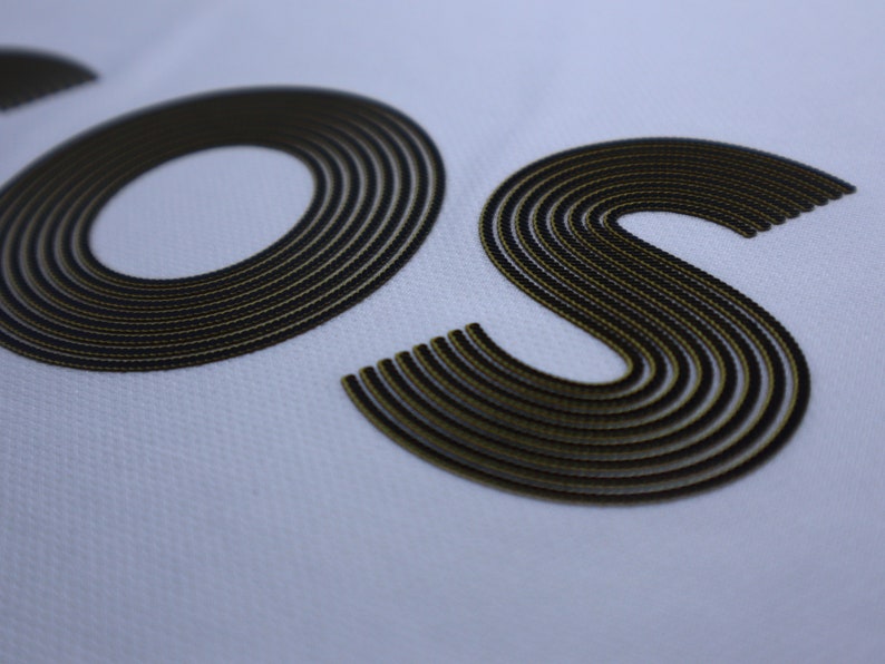 Pode incluir: Close-up das letras "OS" em uma fonte bordada em preto e dourado sobre um fundo branco.