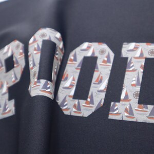 May include: A navy blue shirt with the word "POOL" printed in white letters. The letters are filled with a pattern of small sailboats.