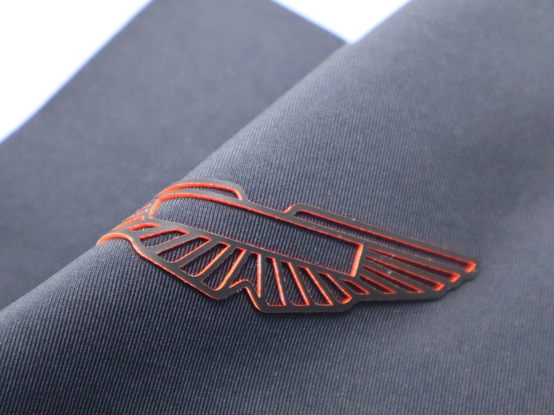 May include: A black and orange winged emblem with a rectangular shape in the center. The emblem is made of a thin, cut-out material and is attached to a dark gray fabric.