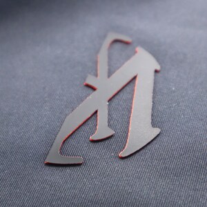 May include: A black letter "A" with a red outline, sewn onto a dark blue fabric.