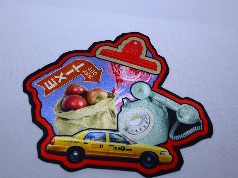 Peut inclure: Un patch color&eacute; avec un panneau de sortie rouge, blanc et bleu, un sac de pommes rouges, un t&eacute;l&eacute;phone rotatif vert vintage et un taxi jaune. Le patch est entour&eacute; d'une bordure noire et d'un contour rouge.
