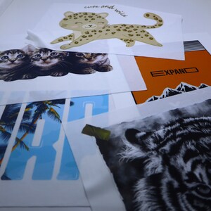 May include: A collection of iron-on transfers featuring various animal designs. The transfers include a leopard, a tiger, and three kittens. The transfers are on white paper and are ready to be applied to fabric.