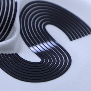 May include: Close-up of a white fabric with a black, raised, S-shaped design. The design is made up of multiple, thin, parallel lines.