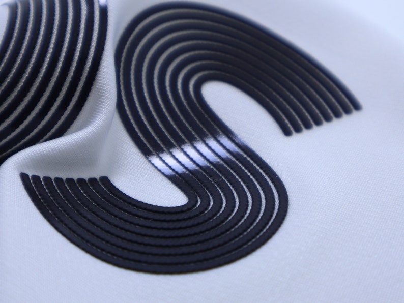 May include: Close-up of a white fabric with a black, raised, S-shaped design. The design is made up of multiple, thin, parallel lines.