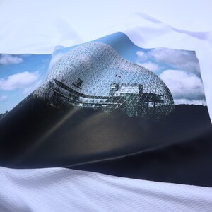 May include: A white t-shirt with a printed image of a large, geodesic dome structure against a blue sky with white clouds. The dome is made of a latticework of metal and glass.
