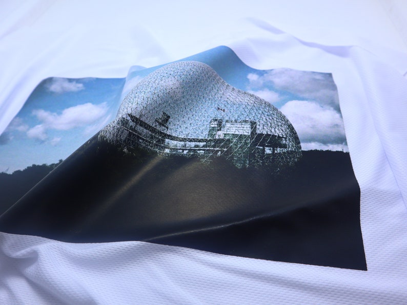 May include: A white t-shirt with a printed image of a large, geodesic dome structure against a blue sky with white clouds. The dome is made of a latticework of metal and glass.