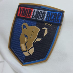 May include: A white, blue, and red patch with the text "YOUR LOGO HERE" on a white background. The patch is attached to a gold and blue shield with a stylized lion head design.