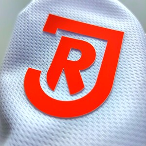 Può includere: Primo piano di un tessuto bianco strutturato con un grande logo arancione brillante. Il logo presenta le lettere "J" e "R" in un design stilizzato e moderno. Il tessuto ha un motivo sottile e ripetitivo.
