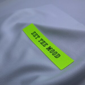 May include: A bright green label with the text "SET THE MOOD" in black outlined letters, placed on a white fabric background.